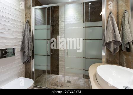 Badezimmer in modernem Stil mit strukturierten Fliesen auf dem Boden und an den Wänden. Zwei Duschen mit Glastür, weiße Toilette und Handtücher. Stockfoto