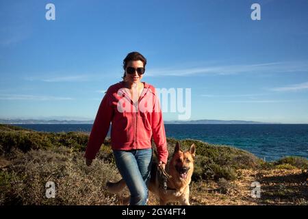 Fröhliche Frau und Hund, die bei Sonnenuntergang an der Küste entlang spazieren. Junge Frau, die ihren Hund trainiert Stockfoto