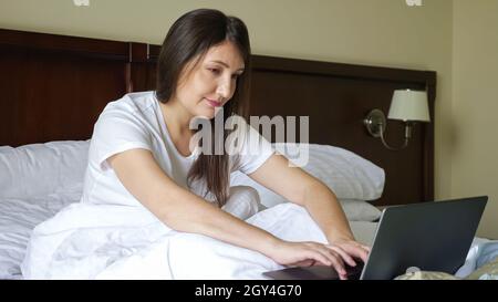 Eine junge Frau tippt auf einem Laptop, während sie im Bett sitzt, das mit einer Decke bedeckt ist Stockfoto