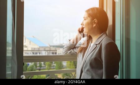 Frau im Geschäftsanzug, die an einem sonnigen Tag am offenen Fenster telefoniert Stockfoto