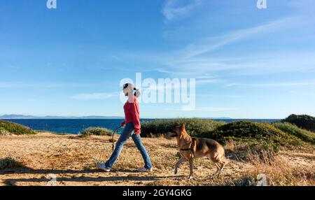 Gesunde, fit junge Frau trainiert ihren Schäferhund, während sie gemeinsam in der Frühlingssonne über Küstensanddünen mit Meer b spazieren gehen Stockfoto