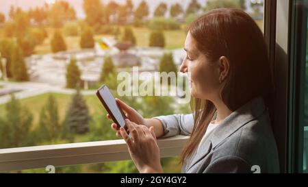 Frau im Geschäftsanzug tippt am Telefon am offenen Fenster Stockfoto