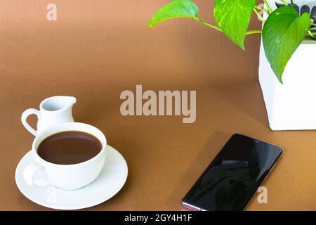 Kaffee mit Milchkännchen auf braunem Hintergrund. In der Nähe gab es ein Handy. Und es gibt eine Topfpflanze in der Ecke des Tisches. Stockfoto