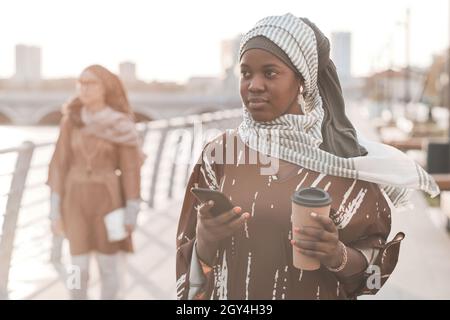Afrikanische muslimische Frau trinkt Kaffee und benutzt Handy während ihres Spaziergangs in der Stadt Stockfoto