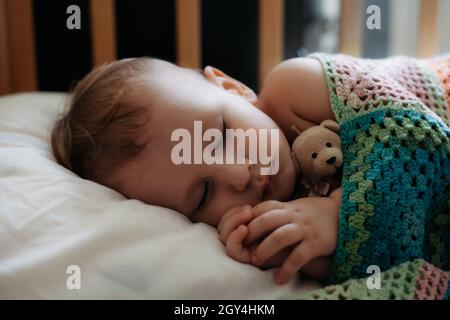 Niedliches Baby schläft friedlich in der Krippe mit Griff unter seiner Wange. Ein Jahr alter Junge liegt bedeckt mit DIY Strickdecke Stockfoto