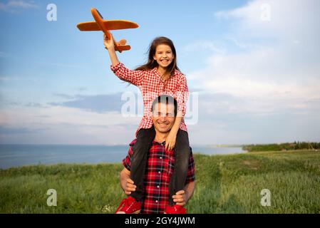 Vater hält sich auf den Schultern ein kleines Mädchen, das seine Tochter vor dem Hintergrund des Himmels mit einem Modellflugzeug spielt, läuft auf dem Feld. Stockfoto