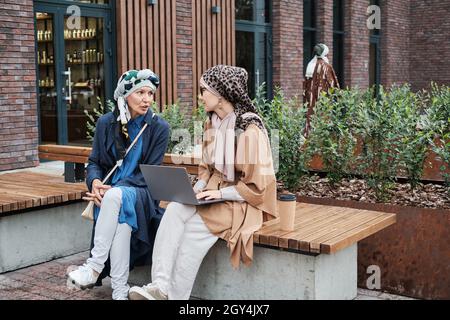 Junge muslimische Frau, die auf der Bank sitzt und auf einem Laptop tippt und draußen mit einer anderen Frau über etwas diskutiert Stockfoto