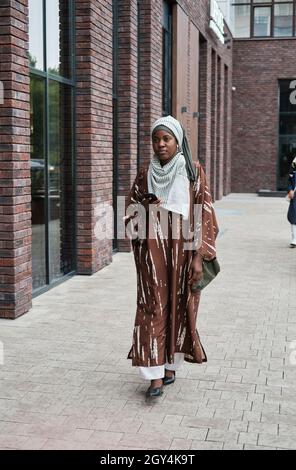 Afrikanische junge Frau in muslimischer Kleidung, die ihr Mobiltelefon hält und draußen auf der Straße entlang geht Stockfoto