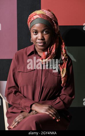 Porträt einer afrikanischen jungen muslimischen Frau im Hijab, die auf einem Stuhl sitzt und die Kamera gegen die moderne Wand anschaut Stockfoto