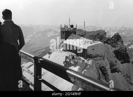 Das Foto zeigt das Münchner Haus auf der Zugspitze um das Jahr 1935, von der Bergstation der Seilbahn zum Schneefernerhaus aus gesehen. Ein Mann schaut auf das Dach der Hütte, das komplett mit Schnee und Eis bedeckt ist. Im Hintergrund erheben sich die Lechtaler Alpen. [Automatisierte Übersetzung] Stockfoto