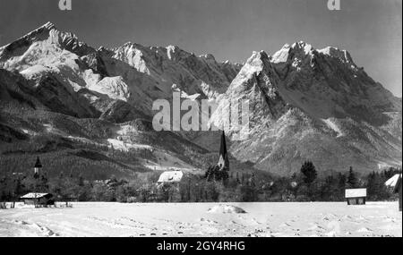Die Kirchtürme der beiden Kirchen im Bezirk Garmisch (St. Martin's Parish Church links, St. Martin's Old Parish Church rechts) steht im winterlich verschneiten Tal vor den eisigen Gipfeln der Hochberge (von links nach rechts: Alpspitze, Höllentalspitzen, Manndl, Waxenstein, Zugspitze). Links von der alten Kirche befindet sich das neue Schulgebäude (Burgstraße 9, heute Grundschule) im barocken Jugendstil. Das Foto aus dem Atelier von Rudolf Rudolphi aus Garmisch wurde im Winter 1927 aufgenommen. [Automatisierte Übersetzung] Stockfoto
