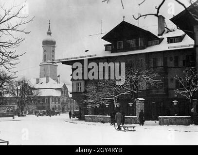 Am 4. Februar begann es in Garmisch-Partenkirchen zu schneien. Gerade rechtzeitig, denn der Schnee war für die Eröffnung der Olympischen Winterspiele am 6. Februar und die folgenden Wettkämpfe notwendig. Auf dem Bild zieht ein Mann seinen großen Schlitten durch den frischen Neuschnee über den Marienplatz, vorbei am Haus Nr. 13-15, in dem sich ein Hotel befand. Links dahinter die alte Apotheke (Hausnr 10) und dahinter ist die Pfarrkirche St. Martin zu sehen. Links hängen Werbeplakate für die Olympischen Spiele an einem Brett. [Automatisierte Übersetzung] Stockfoto