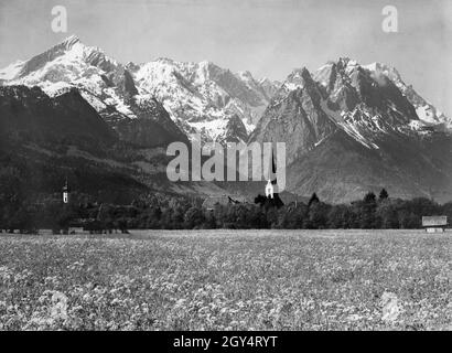 Die Kirchtürme der beiden Kirchen im Bezirk Garmisch (St. Martin's Pfarrkirche links, St. Martin's Alte Pfarrkirche rechts) steht im Frühsommertal mit seinen blühenden Wiesen vor den noch schneebedeckten Gipfeln der hohen Berge (von links nach rechts: Alpspitze, Höllentalspitzen, Manndl, Waxenstein, Zugspitze). Links von der alten Kirche befindet sich das neue Schulgebäude (Burgstraße 9, heute Grundschule) im barocken Jugendstil. Das Foto aus dem Atelier von Rudolf Rudolphi aus Garmisch wurde 1928 aufgenommen. [Automatisierte Übersetzung] Stockfoto
