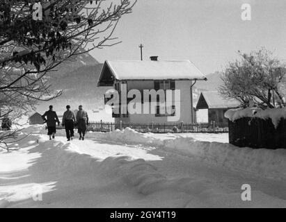 Drei Männer wandern auf einem gerodeten Weg bei Garmisch-Partenkirchen hinaus in die Winterlandschaft. Auf der rechten Seite ist ein kleines Landhaus. Das Foto wurde im Februar 1936 während der Olympischen Winterspiele aufgenommen. [Automatisierte Übersetzung] Stockfoto