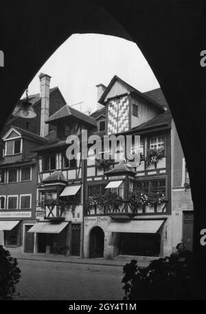 'Das Bild wurde unter einer Arkade des Gasthauses zum Sünfzen aufgenommen und zeigt alte Fachwerkhäuser in der Maximilianstraße in Lindau. Über dem Eingang zum Haus am alten Markt Nr. 4 (Mitte des Bildes) steht ''Hermann Rupflins''. Links davon befindet sich im Haus Nr. 6 ein Friseur. Ganz links im Haus Nr. 8 befand sich im Erdgeschoss das ehemalige Schweizer Getreidehandhaus, die ''Feinbäckerei Ernst H[...]''. Foto undatiert, vermutlich um 1925 aufgenommen. [Automatisierte Übersetzung]' Stockfoto