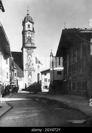 'Der Blick vom Obermarkt geht auf die Pfarrkirche St. Peter und Paul in der Matthias-Klotz-Straße, deren Turm mit Fresken der beiden Namensvetter bemalt ist. Am Eckhaus rechts zur Hochstraße zeigt ein Schild in Richtung Parkenkirchen und Walchensee. Vorne rechts befindet sich das Gasthaus z. Alpenrose v. Ludwig Schweiger''. Auf der linken Straßenseite ist ein Bus zu sehen. Das Foto aus dem Atelier von Rudolf Rudolphi aus Garmisch wurde 1922 aufgenommen. [Automatisierte Übersetzung]' Stockfoto