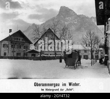 „die Pferde können sich nur mit einem Schlittenwagen durch den Oberammergau, der im Winter 1921 tief im Schnee liegt, durchfahren lassen. Links zeigt ein Schild nach links in die Schnitzlergasse zur Konditorei von Max Hesse. Im Haus in der Bildmitte befindet sich die Drogerie Ammergau mit einer Foto-Handlung, die Kodaks verkauft, und die Pension weil. Rechts auf dem Bürgersteig der Dorfstraße beugt sich eine Frau vor dem Eingang zu einem Zigarrenladen. Der Kofel ist im Hintergrund zu sehen. [Automatisierte Übersetzung]' Stockfoto