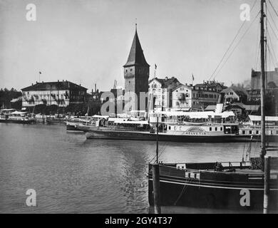 "Schiffe werden im Hafen von Lindau vor dem Mangturm vertäut, der früher als Leuchtturm genutzt wurde. Eines der Passagierschiffe im Hafen ist die ''SD Helvetia III''. An der Seepromenade haben sich Hotels etabliert, darunter das Hotel Bayerischer Hof (links) und das Hotel Reutemann (Mitte) direkt neben Johannes Thomanns Gasthof zur Helvetia. Das undatierte Foto, vermutlich um 1900 aufgenommen, stammt vom Kunsthändler Georg Stuffler in München. [Automatisierte Übersetzung]' Stockfoto