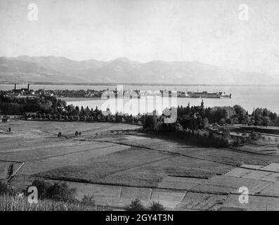 Der Blick geht vom Hoyerberg auf das Wiesental und Bad Schachen (im Vordergrund) zur Insel Lindau mit der Altstadt und dem Bahnhof im Jahr 1903. Der Turm der Villa Wacker in der Schachener Straße 53 erhebt sich aus dem Waldgebiet am Ufer des Bodensees. Im Hintergrund sind die Vorarlberger Alpen im Dunst zu sehen. [Automatisierte Übersetzung] Stockfoto