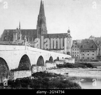 Die Steinbrücke führt über die Donau in die Altstadt von Regensburg. Um 1900 befand sich im Haus rechts von der Brücke das ''Blechwaaren-Geschäft Carl Körper''. 1901/1902 wurde ein Durchgang durch das Haus für größere Fahrzeuge durchtrennt, die nicht durch das Tor unter dem Brückenturm passten. Der Turm wird von Salzstadel (links) und Amberger Stadel (rechts) begrenzt. Der Regensburger Dom ist im Hintergrund. [Automatisierte Übersetzung]' Stockfoto