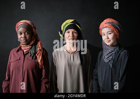 Porträt von drei eleganten Frauen in Hijabs, die die Kamera vor dem schwarzen Hintergrund betrachten Stockfoto