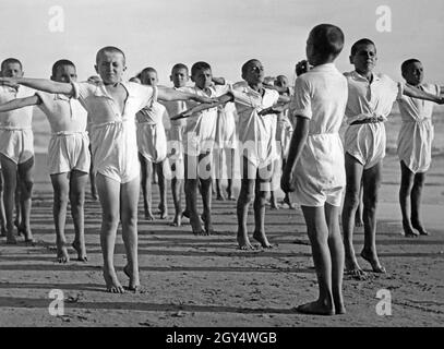 Eine Gruppe von Jungen macht am Strand von Nettuno in Italien Gymnastik, einer von ihnen gibt die Signale und Anweisungen mit einer Pfeife. Auf dem Bild üben sie, ihr Gleichgewicht zu halten, indem sie auf Zehenspitzen stehen und ihre Arme zur Seite strecken. Die Jungen nahmen 1931 am Sommercamp der Opera Nazionale Balilla Teil, das vom faschistischen Föderalen Sekretariat von Rom organisiert wurde. [Automatisierte Übersetzung] Stockfoto