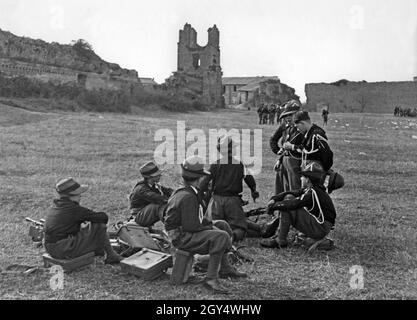 Eine Gruppe von Kindern der Italienischen Oper Nazionale Balilla machen während eines trainingsmarsches 1936 eine Pause auf einer Wiese. Ihre Marschpacks und Rucksäcke dienen den Jungen als Sitze. Zwei von ihnen diskutieren, einer zeigt mit dem Finger auf (wahrscheinlich) ein Gewehr. Im Hintergrund sind Ruinen und ein verlassenes Gehöft zu sehen. [Automatisierte Übersetzung] Stockfoto