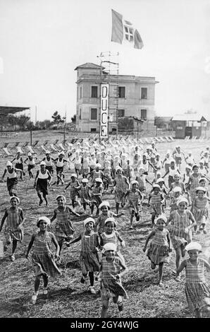 „Dutzende von Schülern laufen auf die Kamera zu. Die Mädchen tragen karierte Kleider, die Jungen Shorts und weiße Hemden. Im Hintergrund befinden sich Liegestühle und eine italienische Flagge fliegt auf einem Gerüst mit der Aufschrift ''DUCE''. Das Bild wurde 1934 aufgenommen, vermutlich in einem Ferienlager für die Kinder. [Automatisierte Übersetzung]' Stockfoto