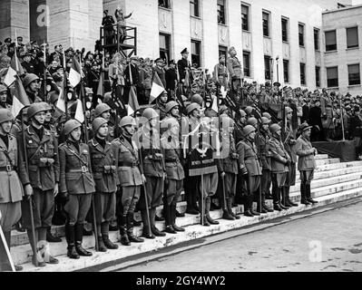 Am 30. Mai 1942 feierte die 'Milizia Universitaria', eine Unterorganisation der Milizia Volontaria per la Sicurezza Nazionale (MVSN) für Studenten, ihr 18-jähriges Bestehen. Die Feier fand in der Città universitaria in Rom statt. Auf den Stufen des Palazzo del Rettorato steht Benito Mussolini, hochklatschig, umgeben von Parteiunterstützern (Hintergrund) und Milizionären der 'IV Legione Universitaria Benito Mussolini' (Vordergrund). Ein Dirigent (Hintergrund, stehend auf einer hölzernen Plattform) signalisiert den Teilnehmern der Feier auf der Piazzale della Minerva. [Automatisierte Übersetzung]' Stockfoto