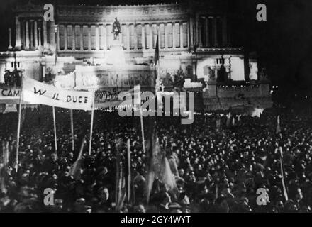 "Am Samstag, dem 9. Mai 1936, wurde das "Italienische Reich" durch die Proklamation von Benito Mussolini gegründet und im Zuge dessen übernahm Viktor Emanuel III., der König von Italien, den Titel des "Imperators von Abessinien". Das faschistische Italien erklärte den Abessinischen Krieg siegreich und alle Gebiete, die der Souveränität Italiens untergeordnet waren. Das Bild zeigt die Menge auf der Piazza Venezia in Rom, die Mussolinis Rede erwartet und vom Balkon des Palazzo Venezia spricht (rechts, nicht im Bild). Im Hintergrund ist der hell erleuchtete Vittoriano zu sehen. Menschen halten Banner hoch, darunter ''VV [Evviva] il Stockfoto