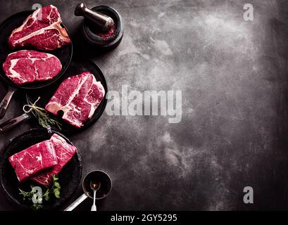 Von oben aus Pfannen mit einem Stück rohem Rindersteak, serviert mit Kräutern und Gewürzen auf einem grauen Betontisch Stockfoto