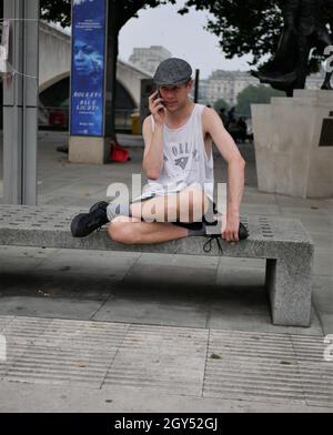 Junger Mann, der am Telefon telefoniert, sitzt auf einer Betonbank entlang der South Bank in London Stockfoto