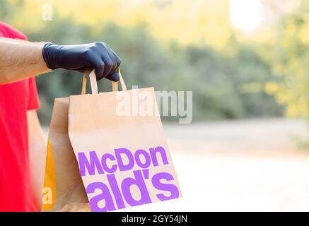 Eine Männerhand in einem medizinischen Handschuh hält eine Papiertasche von Mcdonal. Lieferung McCafe. Fast Food Restaurant McDonalds. Nehmen Sie McDonal's mit Stockfoto