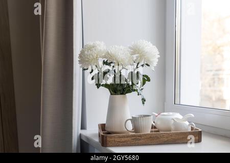 Drei weiße Chrysanthemen in einer gewellten Vase im Stil der 70er Jahre. Holztablett mit Wasserkocher, Milchkännchen und einer Tasse Tee auf der Fensterbank. Frühstück am Morgen Stockfoto