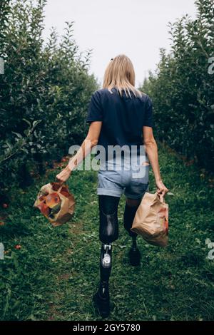 Eine behinderte Frau in Prothesen trägt zwei Papiertüten mit Äpfeln. Stockfoto