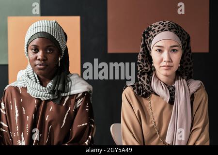 Porträt zweier junger muslimischer Frauen in traditioneller Kleidung, die auf Stühlen mit moderner Wand im Hintergrund die Kamera betrachten Stockfoto