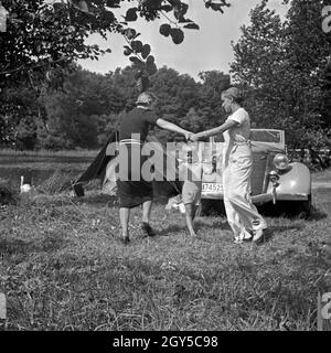 Zwei junge Frauen tanzen Ringelreihen mit einem kleinen Jungen vor einem Ford V8 Cabrio am Ufer eines sieht, Deutschland 1930er Jahre. Zwei junge Frauen tanzen Ring-a-ring-a-Rosen wit Ha kleine Junge vor einem Ford V8 Cabriolet, Deutschland 1930. Stockfoto