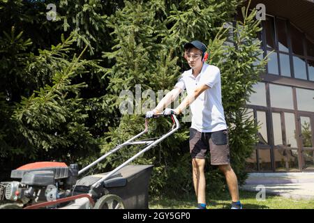 Junger Mann mäht einen Rasen in der Nähe eines großen Herrenhauses. Arbeiter mit einem Benzinrasenmäher Stockfoto
