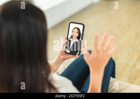 Junge Frau, die auf dem Sofa sitzt und Mobiltelefon hält, Videoanruf täte und mit Kollegen des Unternehmens über Telearbeit diskutierte Stockfoto