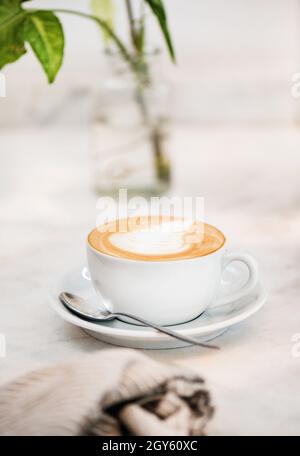 Cappuccino-Kaffeegetränk in weißer Tasse mit Latte Art auf der Oberseite und Löffel über weißem Marmorhintergrund, Draufsicht, selektiver Fokus. Idyllisches Getränk für eine Pause Stockfoto