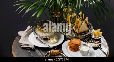Schwarzer Kaviar in Dose auf Eis in silberner Schale, Champagner und Mini-Pfannkuchen auf schwarzem Holzhintergrund Stockfoto