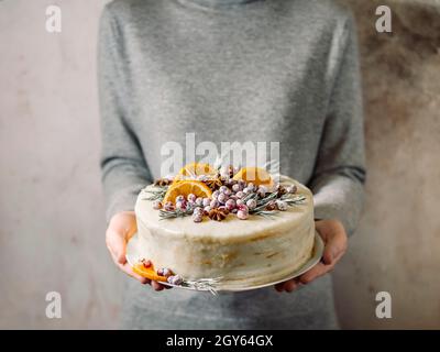 Winter hausgemachter Kuchen in weiblichen Händen. Nicht erkennbare Frau hält weiße Creme weihnachtskuchen dekorierten Rosmarin, Preiselbeeren und Orangen Scheiben Stockfoto