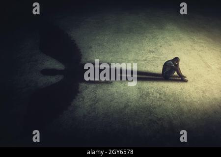 Deprimierter junger Mann, der allein in einem dunklen Raum sitzt und einen Schatten eines mächtigen Schutzengels wirft. Innere Stärke konzeptioneller Hintergrund. Stress Stockfoto