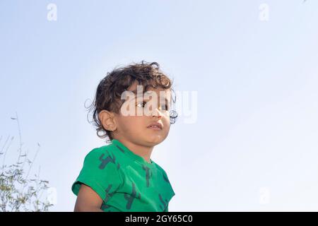 Nahaufnahme Porträt aus dem niedrigen Winkel eines indischen schönen kleinen hindu-Jungen, der draußen vor dem Himmel mit einem grünen T-Shirt posiert Stockfoto