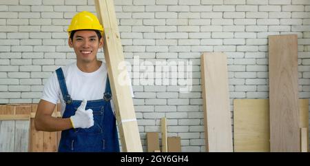 Junger asiatischer Tischler mit gelbem Hut, der Holz für die Möbelherstellung im Werkstattraum trägt. Stockfoto