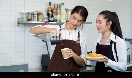 Der junge kaukasische Barista gießt Milch aus dem Krug in eine Papierkaffeebecher. Asiatische Assistentin in einer Schürze, die einen Croissantteller hält, steht neben ihr. Morn Stockfoto