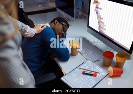 Frau schaut auf schlafende Managerin, Nachtbüro Lebensstil. Müde männliche Personen am Laptop, dunkles Interieur im Hintergrund, moderner Arbeitsplatz Stockfoto