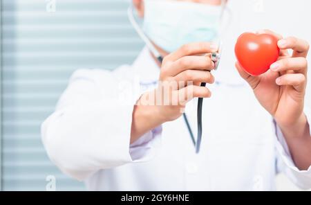 Nahaufnahme eines Arztes, der einen weißen Mantel trägt und sein Stethoskop zur Hand hält, um das rote Herz zu untersuchen und medizinische Geräte zu zeigen Stockfoto