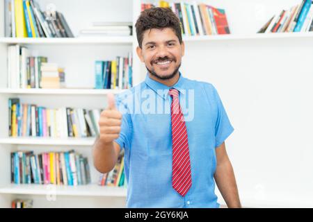 Hispanische reife Geschäftsmann mit Bart und Krawatte zeigt Daumen in Innenräumen im Büro Stockfoto
