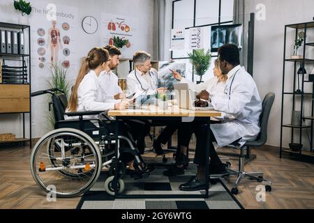 Eine Gruppe von fünf multiethnischen Medizinern, die die Ergebnisse eines Röntgenscans diskutieren, während sie im Büroraum sitzen. Qualifizierte Ärzte und ihre Kollegen im Rollstuhl, die sich in Innenräumen treffen. Stockfoto